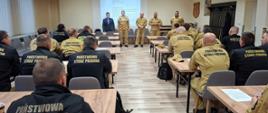 Strażacy stoją w rzędzie mają ubrane piaskowe mundury, obok nich stoi pan w garniturze. Z tyłu widać wyświetloną projekcje na ścianie. Wszystkie osoby znajdują się na sali w której rozmieszczone są ławki przy których siedzą strażacy biorący udział w szkoleniu
