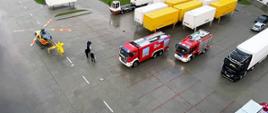 Widać dwa samochody straży pożarnej oraz helikopter lotniczego pogotowia ratunkowe na placu przy hali magazynowej.