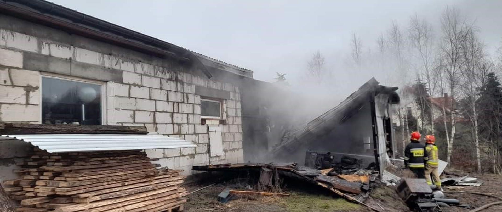 Przed budynkiem gospodarczym i palącą się jego częścią stoi dwóch strażaków w strojach specjalnych i gaszą pożar.