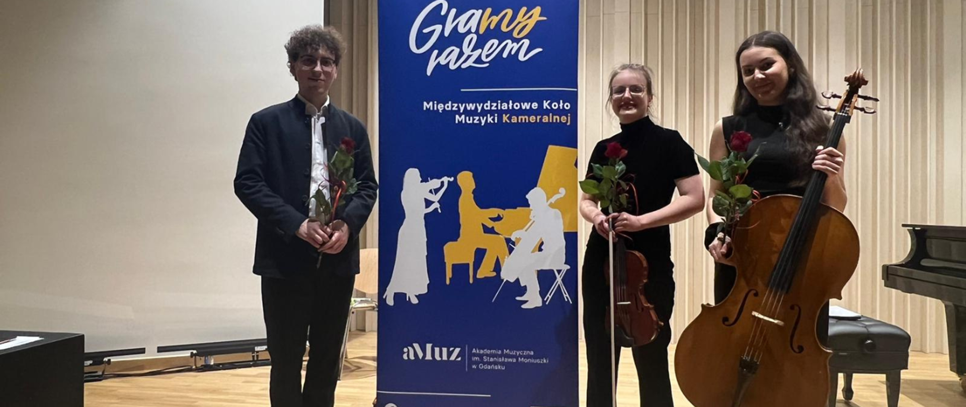 Na zdjęciu po lewej stronie mężczyzna w białej koszuli, czarnych spodniach i granatowej marynarce, nosi okulary i ma bujne loki na głowie. Trzyma w rękach czerwoną różę. Dalej do prawej baner w odcieniach niebieskich z napisem gramy razem, Międzywydziałowe Koło Muzyki Kameralnej. Baner należy do Akademii Muzycznej w Gdańsku. Obok niego po prawej stronie kobieta ubrana na czarna w okularach z czerwoną różą. Na samym końcu brunetka ubrana na czarno, z długimi włosami, czerwoną różą i wiolonczelą. 