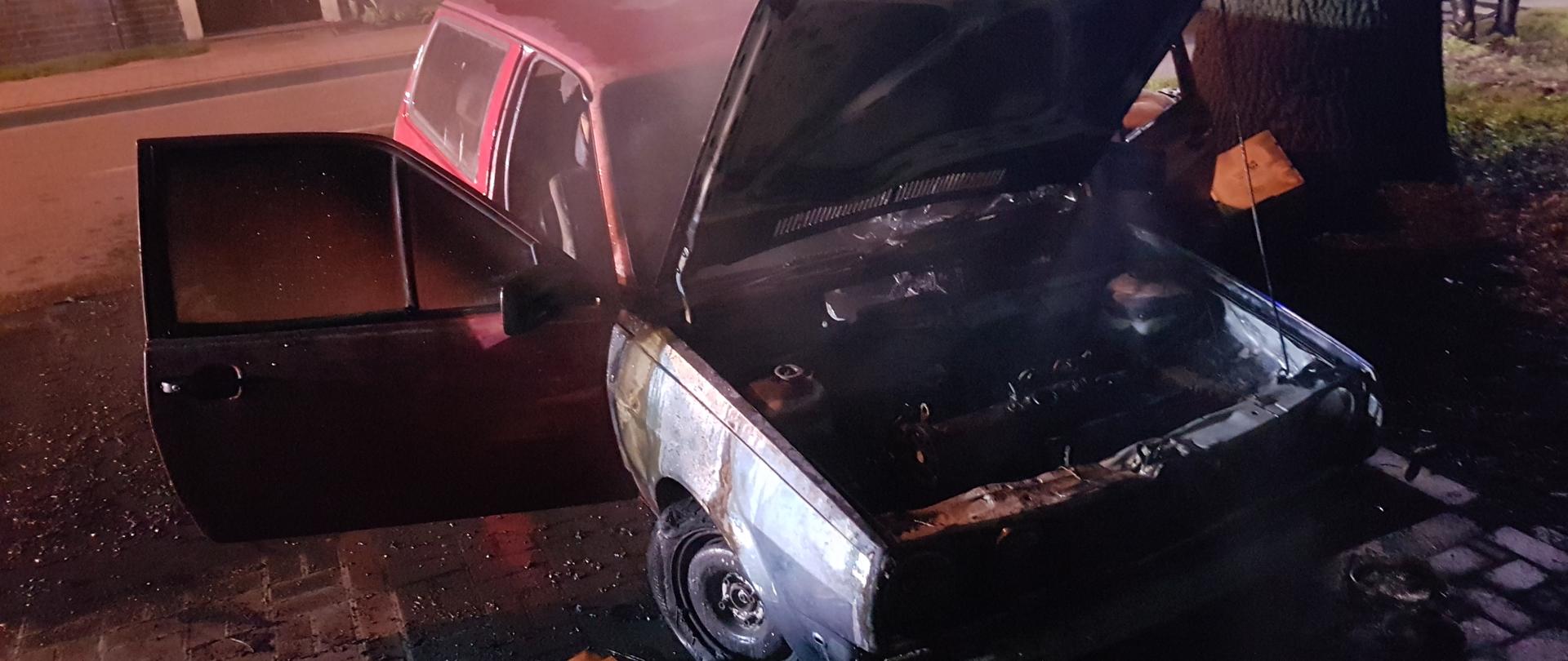 Na zdjęciu spalony samochód osobowy stoi na parkingu. Z tyłu budynek. Pora nocna