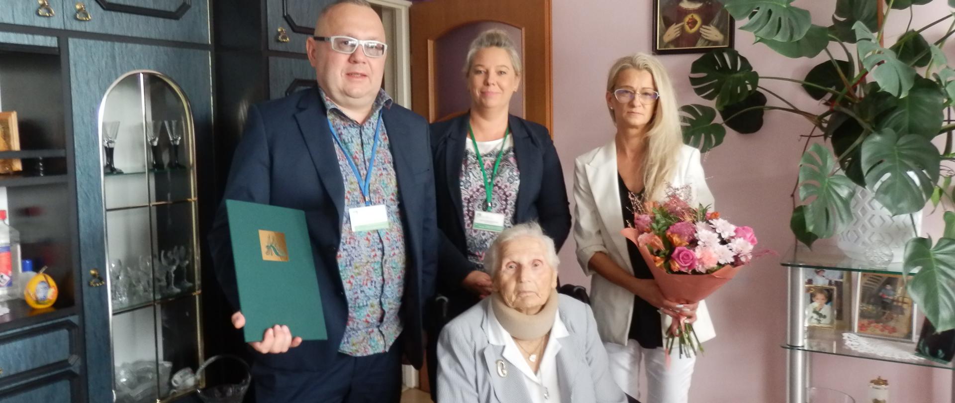 Przy stole na wózku inwalidzkim siedzi Jubilatka – starsza siwa kobieta w szarej marynarce z kocem na kolanach. Za nią stoją: mężczyzna w okularach w granatowym garniturze trzymający w ręce dyplom, kobieta w granatowej marynarce, kobieta w białej marynarce i spodniach trzymająca bukiet różowych kwiatów.