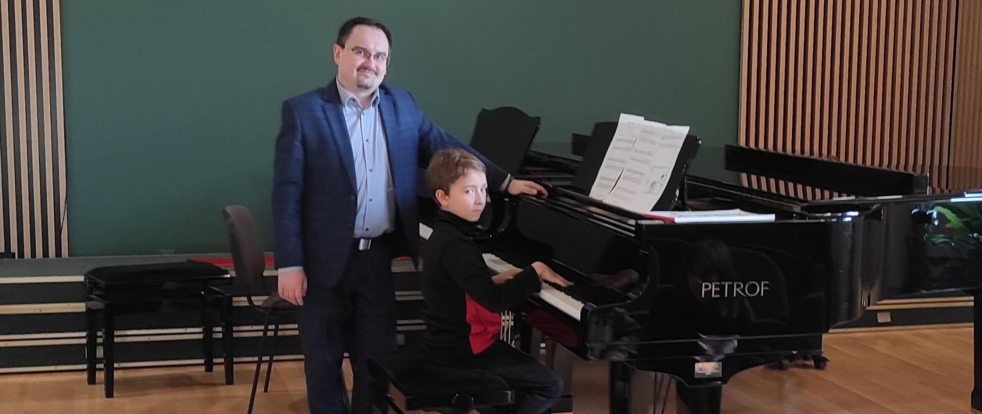 Na zdjęciu widać 2 fortepiany na scenie koncertowej przy pierwszym z nich siedzi uczeń a tuż za nim stoi profesor. Oboje patrzą się w kamerę. W tle widoczna jest ściana w kolorze zielonym z drewnianymi lamelami.