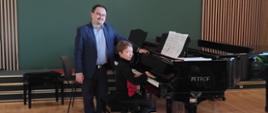 Na zdjęciu widać 2 fortepiany na scenie koncertowej przy pierwszym z nich siedzi uczeń a tuż za nim stoi profesor. Oboje patrzą się w kamerę. W tle widoczna jest ściana w kolorze zielonym z drewnianymi lamelami.