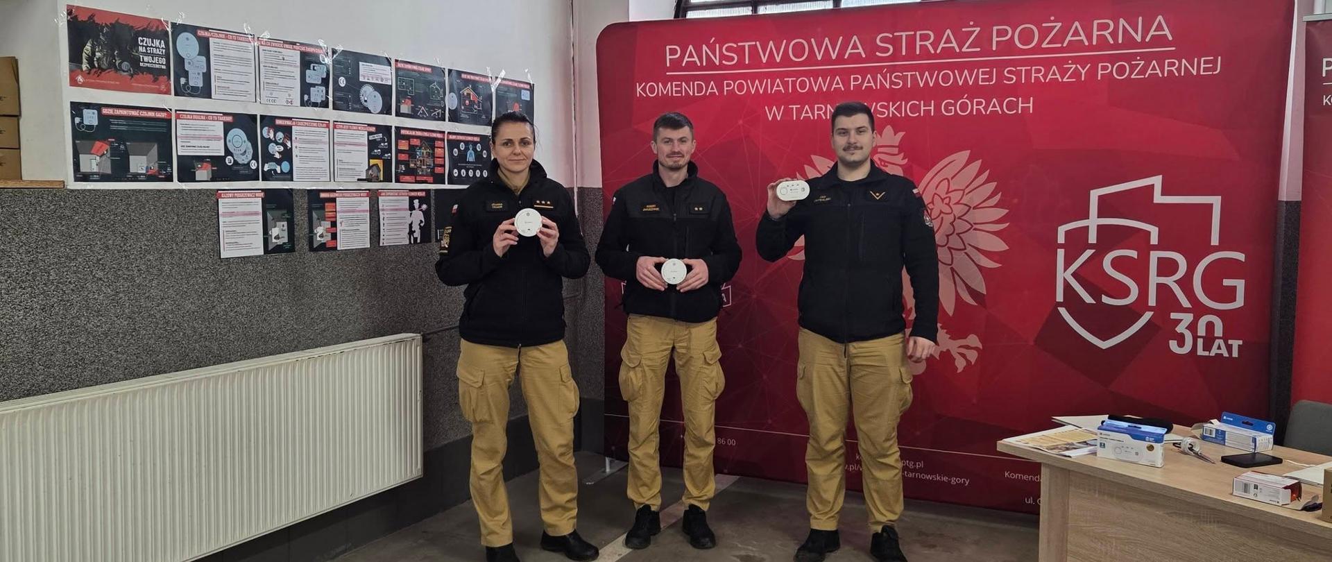 Strażacy: Troje funkcjonariuszy Państwowej Straży Pożarnej (kobieta i dwóch mężczyzn) stoi w rzędzie, prezentując czujki dymu lub tlenku węgla. Są ubrani w pełne mundury służbowe: czarne polary/bluzy oraz charakterystyczne piaskowe spodnie.
Tło i Baner: Za nimi znajduje się duża, czerwona ścianka reklamowa z napisem „PAŃSTWOWA STRAŻ POŻARNA Komenda Powiatowa Państwowej Straży Pożarnej w Tarnowskich Górach”. Po prawej stronie widoczne jest logo KSRG (Krajowy System Ratowniczo-Gaśniczy) z dopiskiem „30 lat”.
Materiały edukacyjne: Na ścianie po lewej stronie widać gazetkę z licznymi infografikami i plakatami. Jeden z nich ma wyraźny tytuł „Czujka na straży Twojego bezpieczeństwa”. Materiały te prawdopodobnie instruują, jak unikać zatrucia czadem i gdzie najlepiej montować urządzenia alarmowe.
Otoczenie: Po prawej stronie stoi biurko, na którym leżą ulotki oraz rozpakowane pudełka z czujnikami, co sugeruje punkt informacyjny, w którym mieszkańcy mogą zasięgnąć rady lub otrzymać takie urządzenie.