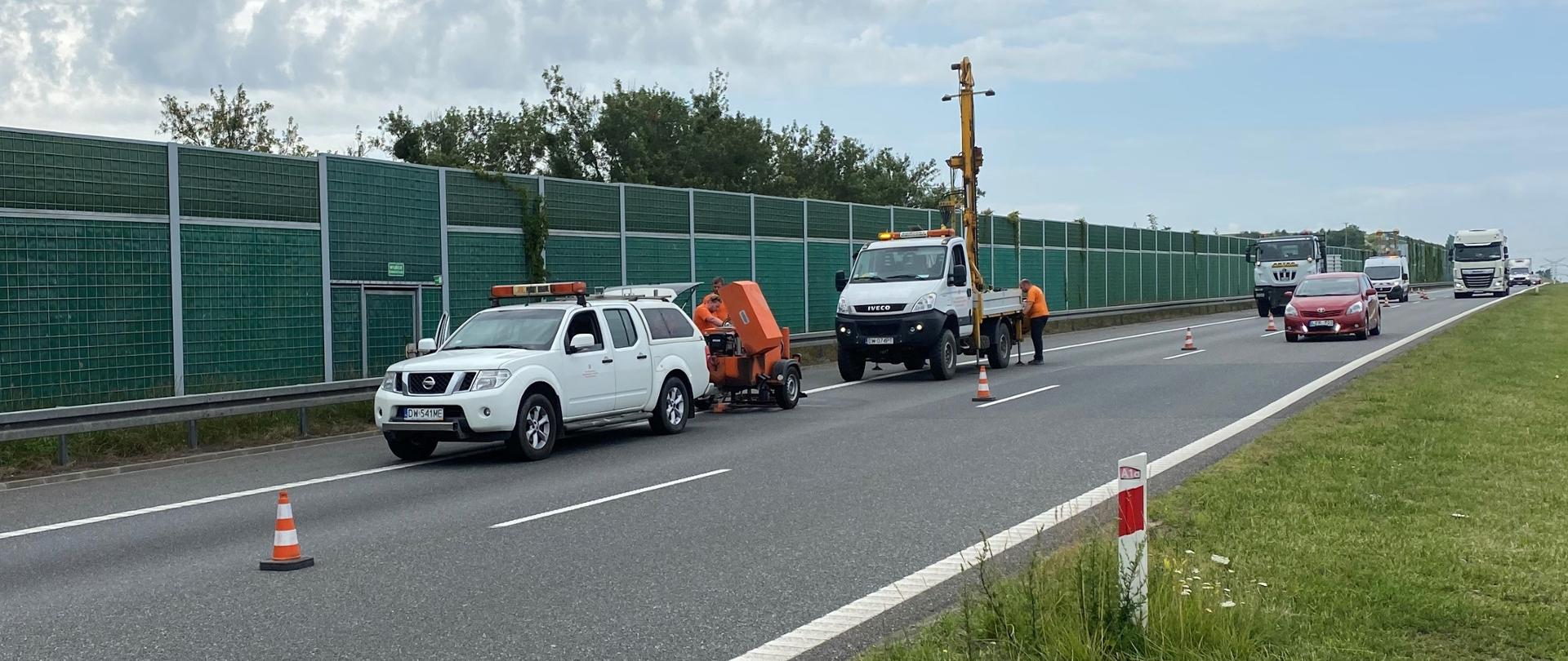 Fotografia obrazująca wykonywanie badań na jezdni autostrady. Widoczne pojazdy specjalistyczne do odwiertów w konstrukcji jezdni wraz z obsługującymi je operatorami i laborantami. Miejsce badań na prawym pasie jezdni wyłączone z ruchu i zabezpieczone słupkami drogowymi. Po lewym pasie poruszają się pojazdy korzystające z autostrady. Autostrada wyposażona jednostronnie w ekrany akustyczne. 