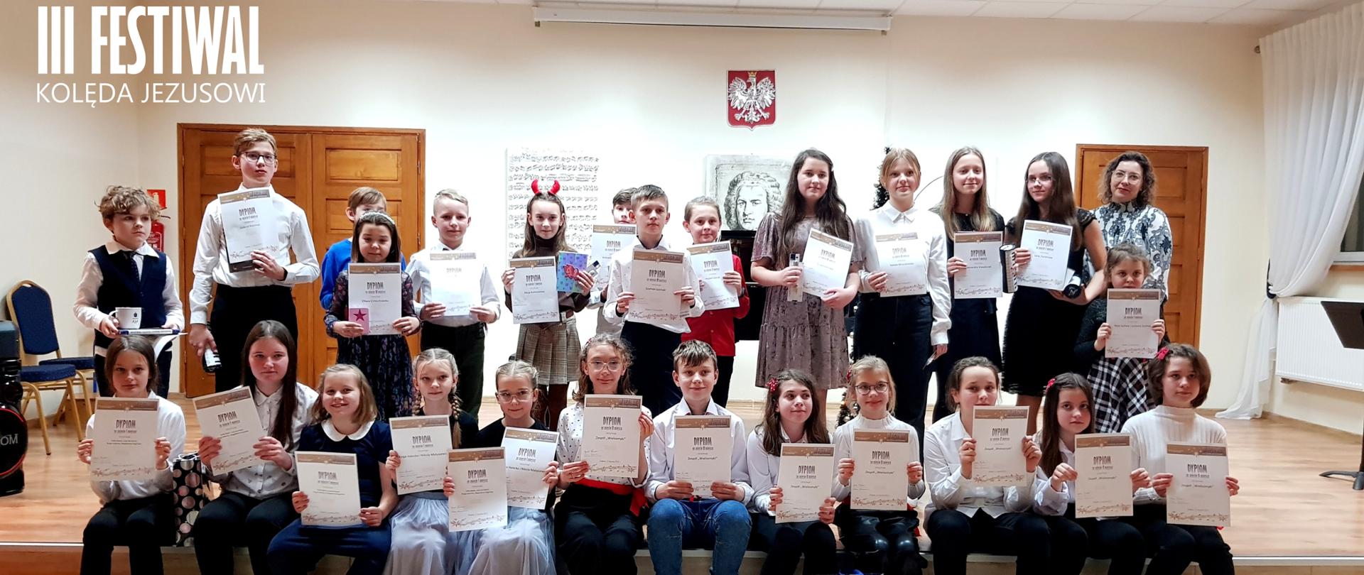Na scenie stoją ustawieni w dwóch rzędach laureaci III Festiwalu "Kolęda Jezusowi", w dłoniach trzymają dyplomy i nagrody. W tle brązowe drzwi, godło polskie, grafika partytury oraz portret Jana Sebastian Bacha. 