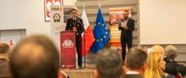 Oficer Państwowej Straży Pożarnej przemawia przy mównicy podczas konferencji w Centralnym Muzeum Pożarnictwa w Mysłowicach. Obok stoi mężczyzna z mikrofonem i kartką. W tle flagi Polski i Unii Europejskiej oraz banery organizatorów wydarzenia.