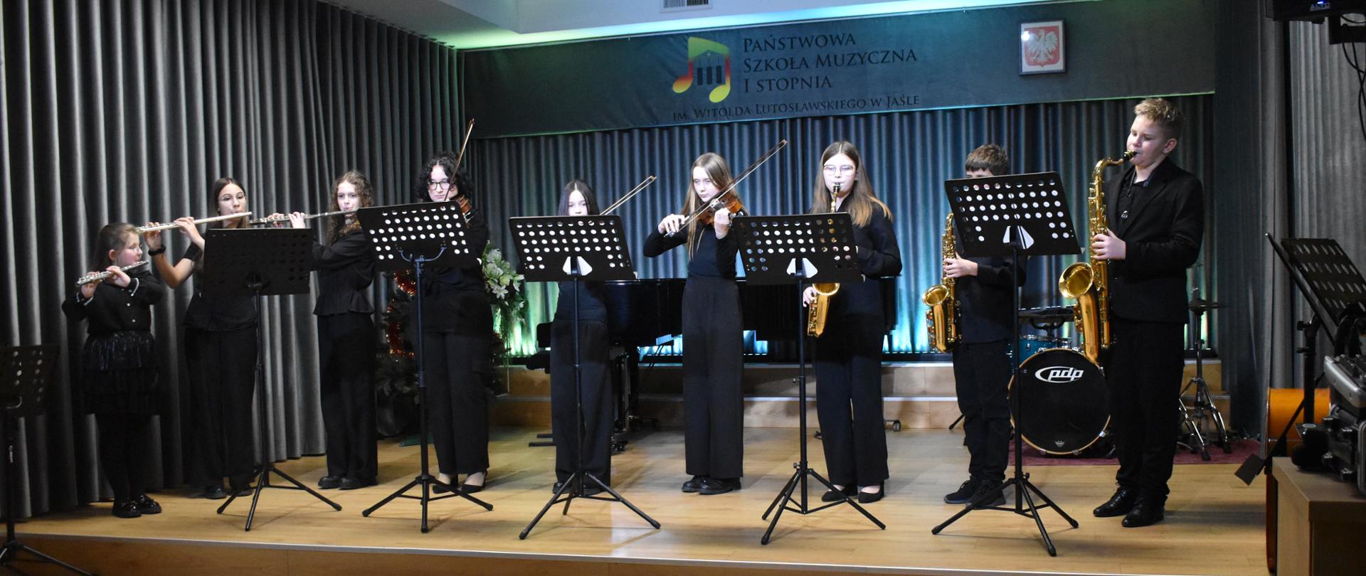 Zespół uczniów grających na różnych instrumentach przed nimi stoją pulpity z nutami. Od lewej strony stoją trzy dziewczynki grające na fletach, kolejne trzy dziewczynki grają na skrzypcach i trójka dzieci z prawej strony gra na saksofonach. Wszystkie dzieci ubrane są na czarno. Za nimi widać fragment perkusji i fortepianu. W tle na ścianie logo Państwowa Szkoła Muzyczna I stopnia im. Witolda Lutosławskiego w Jaśle i godło państwowe .
