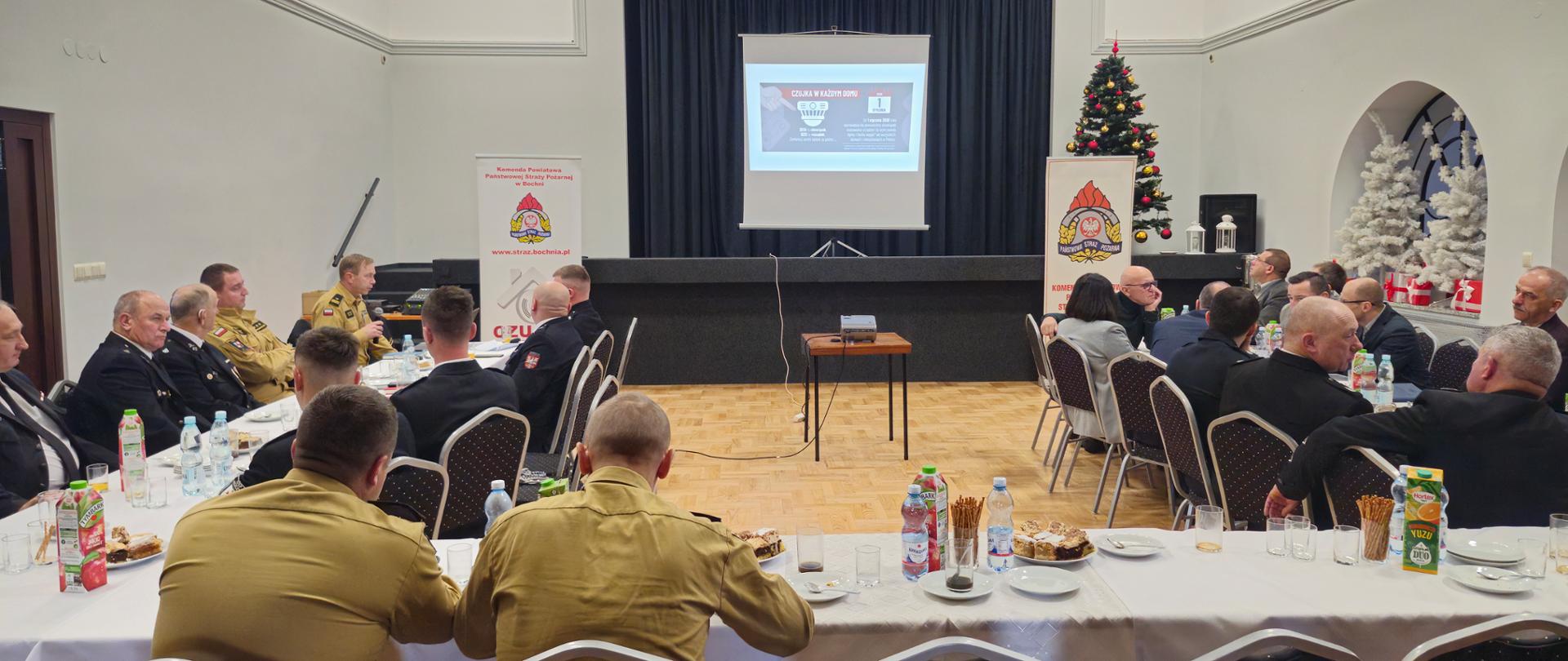Na sali konferencyjnej ze sceną znajdują się grupa strażaków w umundurowaniu służbowym w żółtym kolorze, strażacy w mundurach wyjściowych oraz inne osoby ubrane w stylu każualowym. Jeden ze strażaków przemawia do mikrofonu a na ekranie wyświetlany jest slajd dotyczący czujki dymu.
