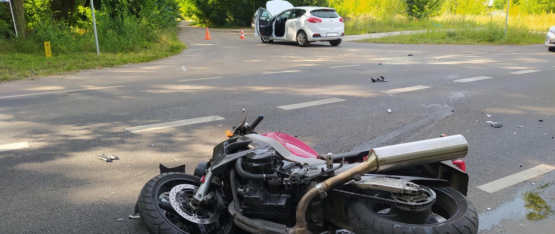 Przewrócony motocykl leży na jezdni. W tle widoczny samochód osobowy.