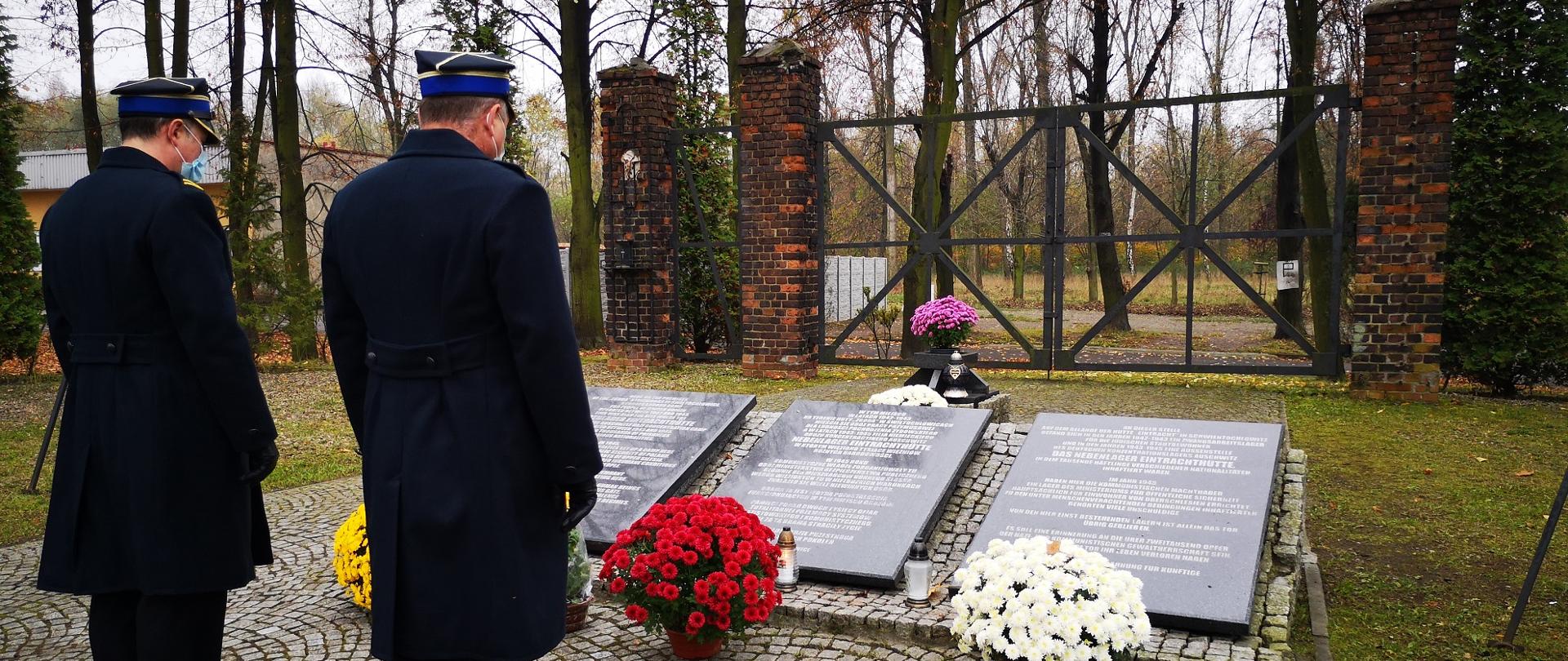 Zdjęcie przedstawia zastępcę komendanta i dowódcę jednostki ratowniczo-gaśniczej stojących przed pomnikiem przy bramie byłego obozu Zgoda.