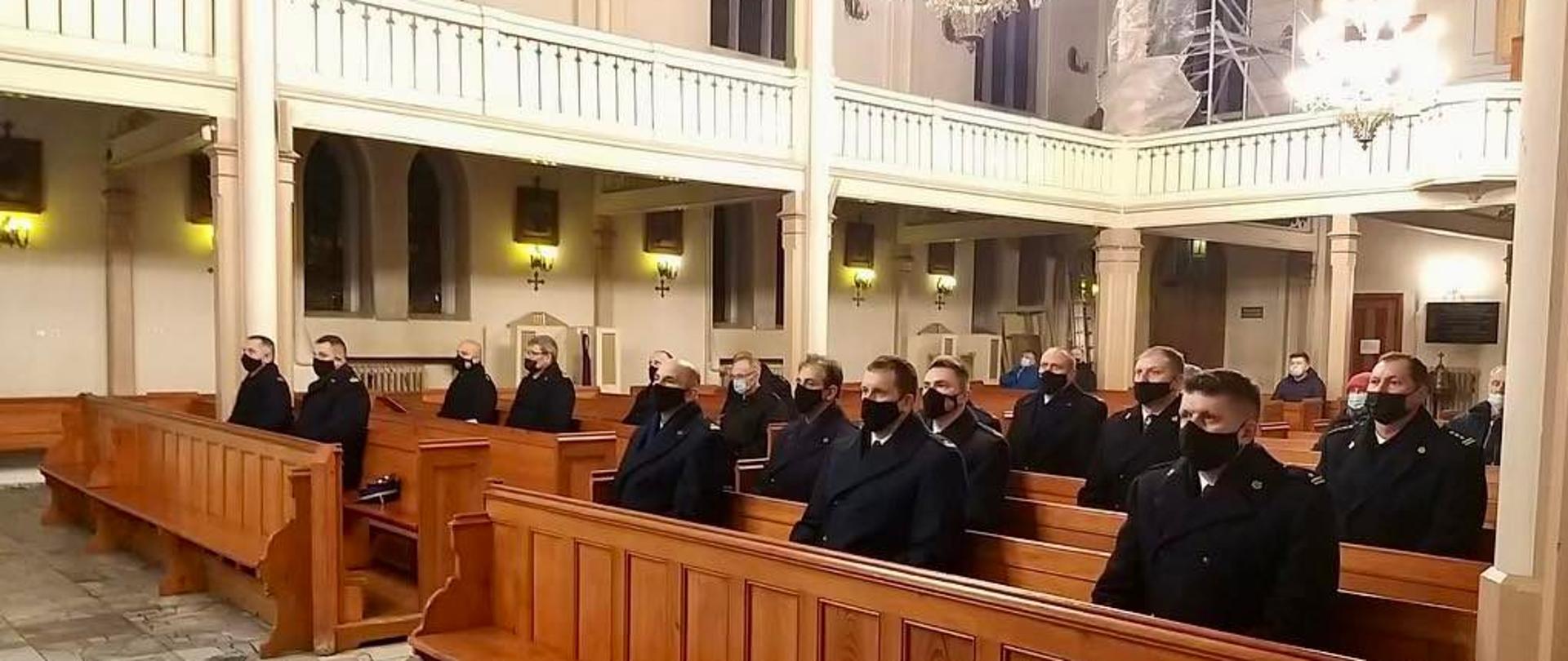 Uczestnicy mszy św. w intencji zmarłych strażaków, w pierwszych drewnianych lawkach w odstępach od siebie siedzą komendanci miejscy/powiatowi Państwowej Straży Pożarnej. W górze widoczne balustrady, w tle widać kawałek organ oraz rusztowanie.