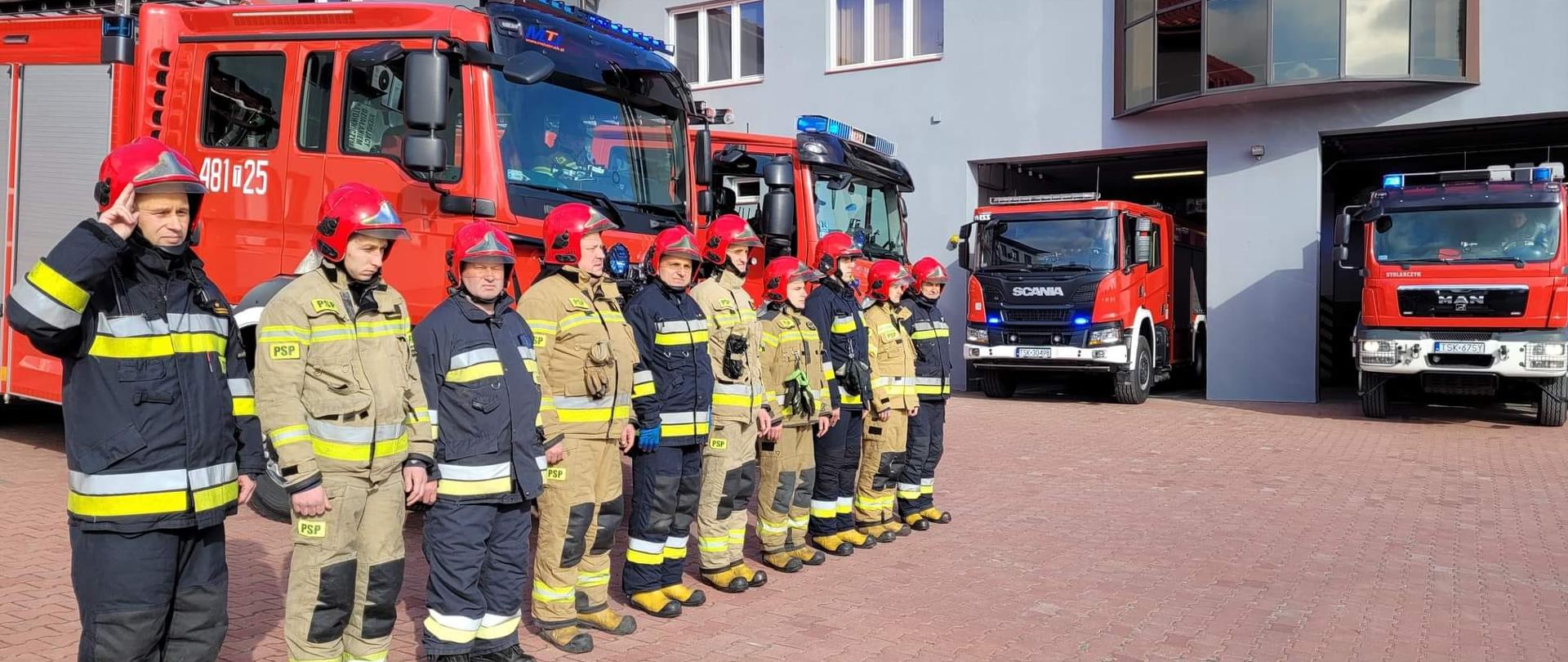 Zdjęcie obrazuje strażaków oddających hołd poległym ofiarom katastrofy smoleńskiej. Strażacy ubrani w ubrania bojowe stoją przed garażami strażnicy . Za nimi stoją wystawione samochody pożarnicze z włączonymi sygnałami świetlnymi.
