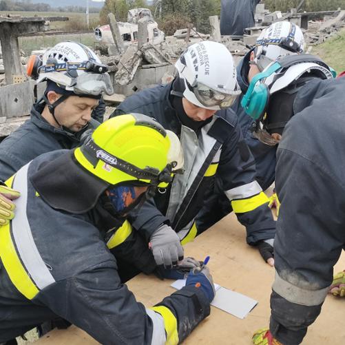 6 strażaków w strojach strażackich i hełmach pochyla się nad stołem, gdzie zapisują coś w notesie. Z tyłu widać zawalony budynek