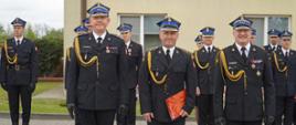 Kujawsko-pomorski Komendant Wojewódzki Państwowej Straży Pożarnej, Komendant Powiatowy Państwowej Straży Pożarnej w Rypinie oraz awansowany na wyższy stopień funkcjonariusz stoją w rzędzie po wręczeniu awansu. W tle ustawiony jest pododdział funkcjonariuszy.