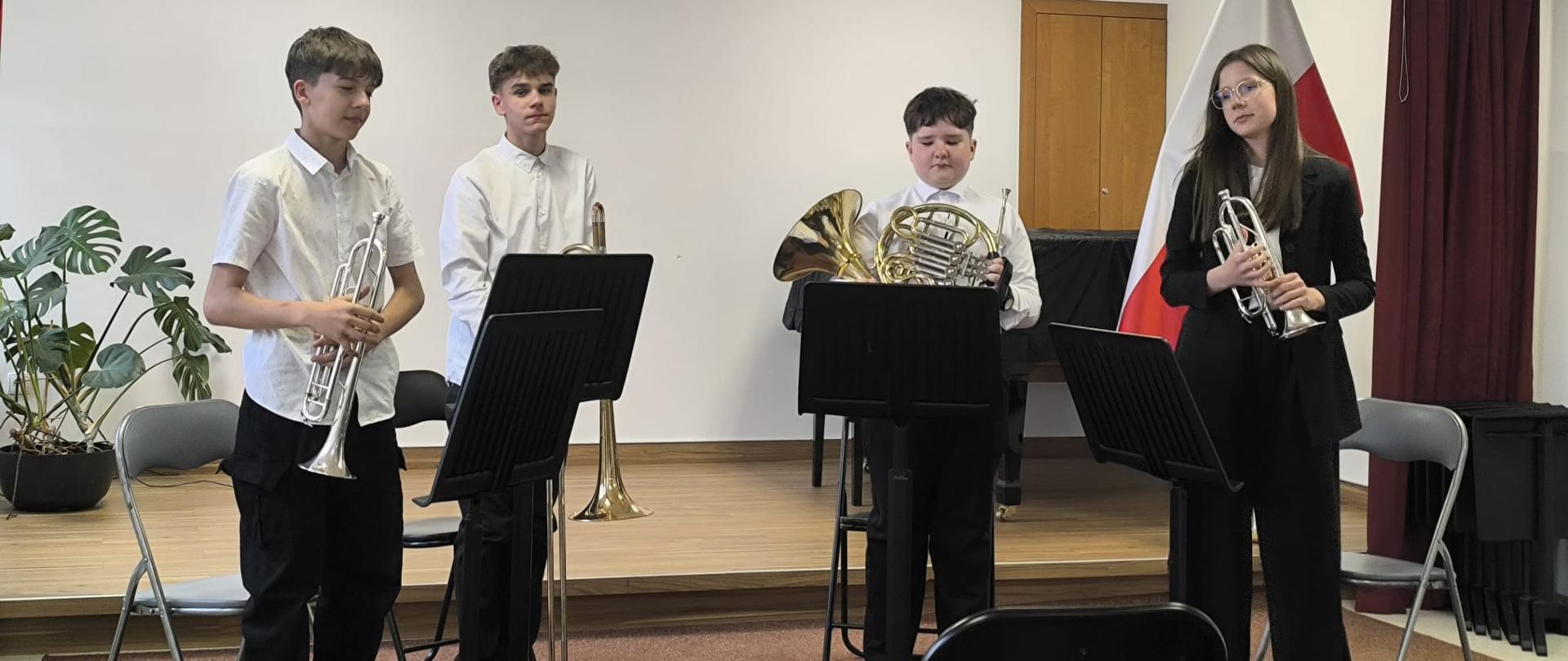 Czterech uczniów z instrumentami dętymi blaszanymi stojących przy pulpitach nutowych podczas występu w V Tuchowskim Konkursie Zespołów Instrumentalnych w Tuchowie.