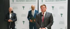 Wiceminister Rzymkowski stoi i mówi do mikrofonu, za nim stoi mężczyzna w niebieskim garniturze i drugi w szarym garniturze, w tle ścianka z napisem Inspekcja Transportu Drogowego.