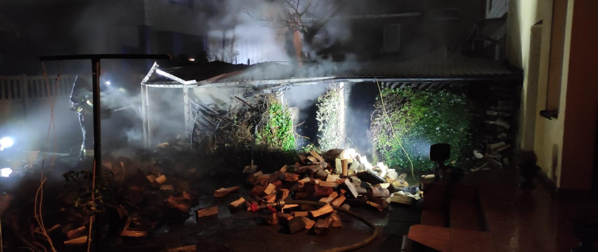 Pożar budynku gospodarczego. Na zdjęciu budynek gospodarczy, unosi się dym, przed porąbane drewno, strażak gasi pożar. Obok budynki mieszkalne.