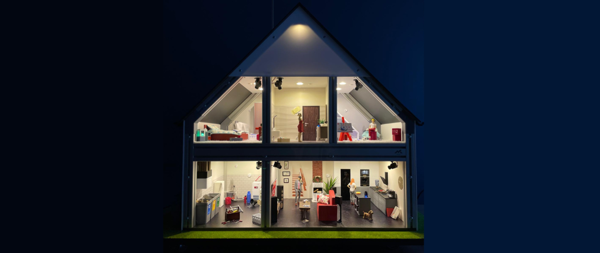 Przekrój nowoczesnego domu pokazany jak domek dla lalek, widoczny nocą na ciemnym tle. Wnętrze jest podzielone na dwa piętra z oświetlonymi pomieszczeniami: kuchnią, salonem, łazienką i sypialnią. W środku znajdują się miniaturowe meble, przedmioty codziennego użytku oraz małe figurki ludzi, co nadaje scenie charakter makiety.