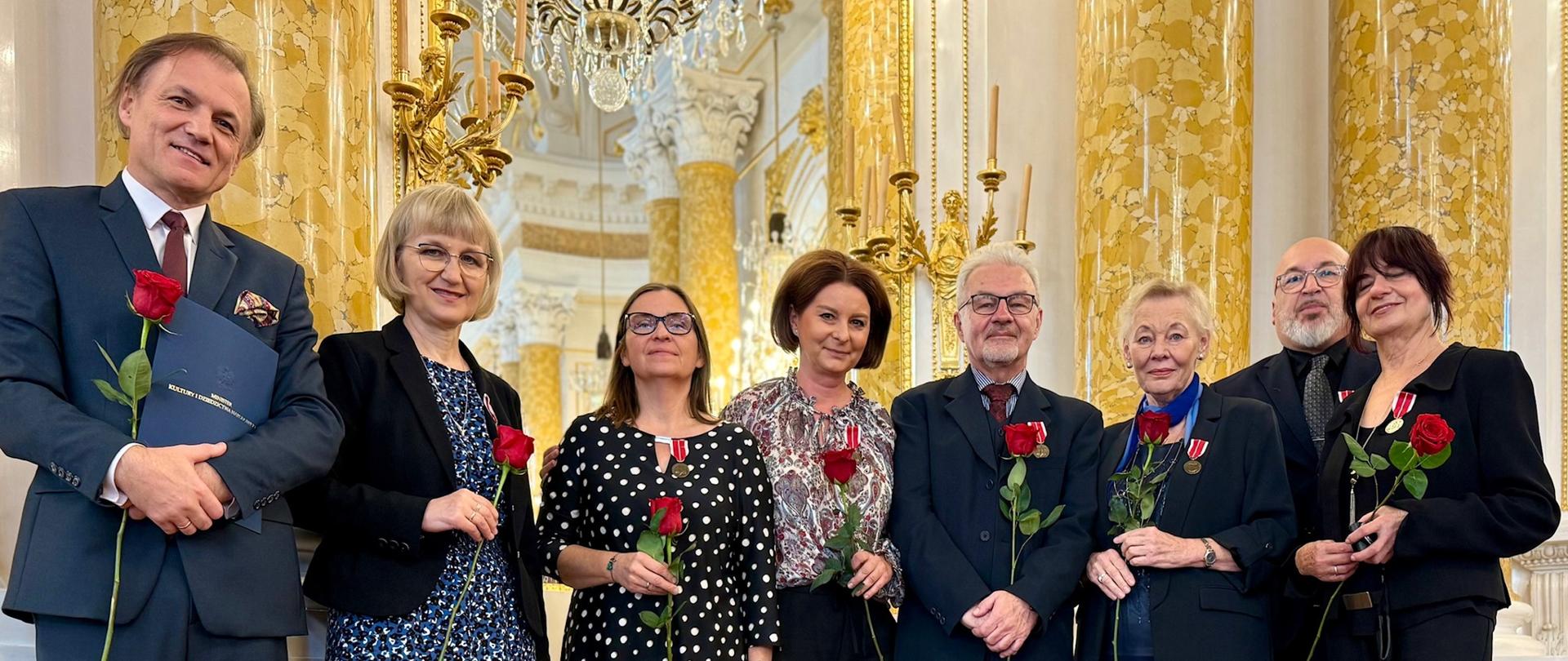 Grupa nauczycieli z odznaczeniami i czerwonymi różami podczas uroczystości wręczenia Medali Komisji Edukacji Narodowej na Zamku Królewskim w Warszawie.