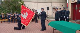 moment nadania sztandaru jednostce osp, jeden druh trzyma pochylony sztandar drugi klęczy na jedno kolano przy płachcie sztandaru, obok lekko w tyle stoi strażak psp oraz 3 druhów, po lewej w tyle orkiestra dęta osp, w tle ściany budynków