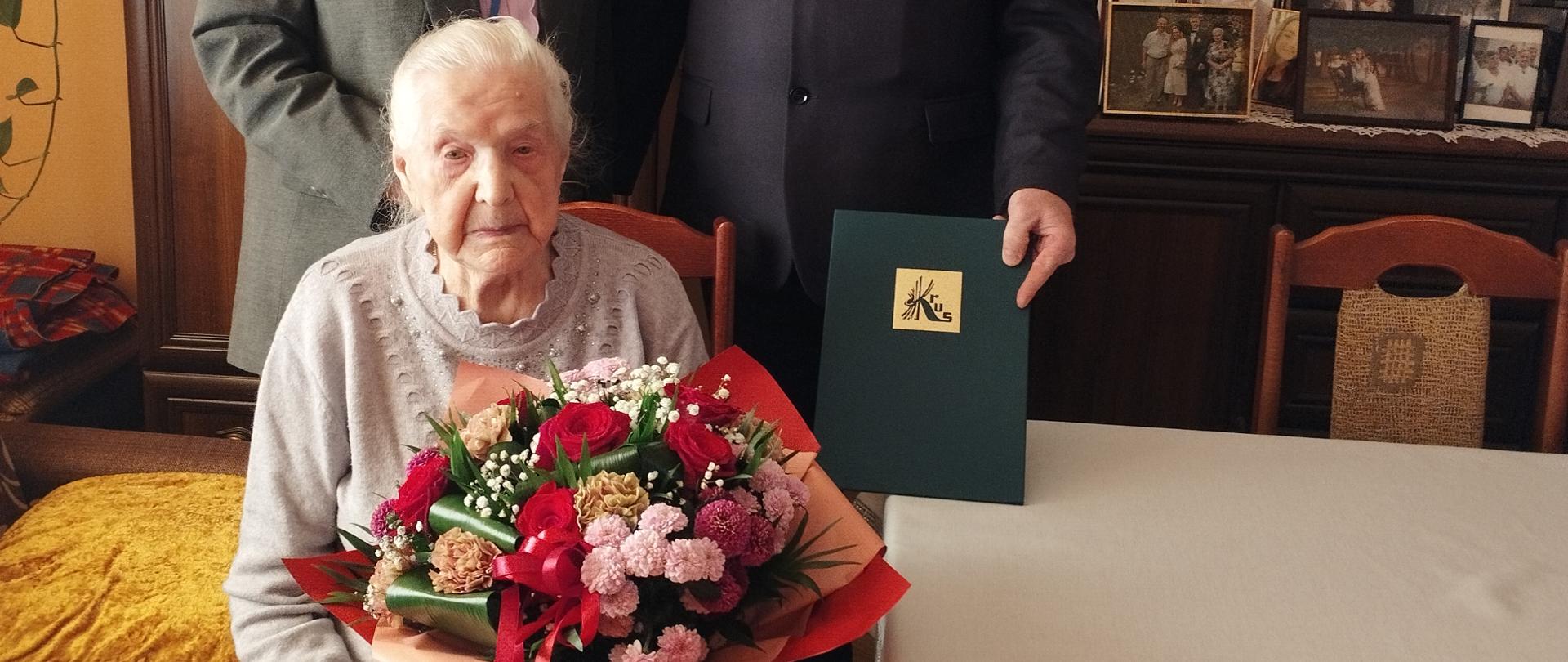 W pokoju obok stołu siedzi na krześle starsza kobieta z bukietem różowo-czerwonych kwiatów, jest przejęta podniosłą chwilą. Za nią stoją mężczyzna i kobieta. Mężczyzna prezentuje ozdobną zieloną teczkę ze złotym logo KRUS. Wszyscy uśmiechają się do zdjęcia.
