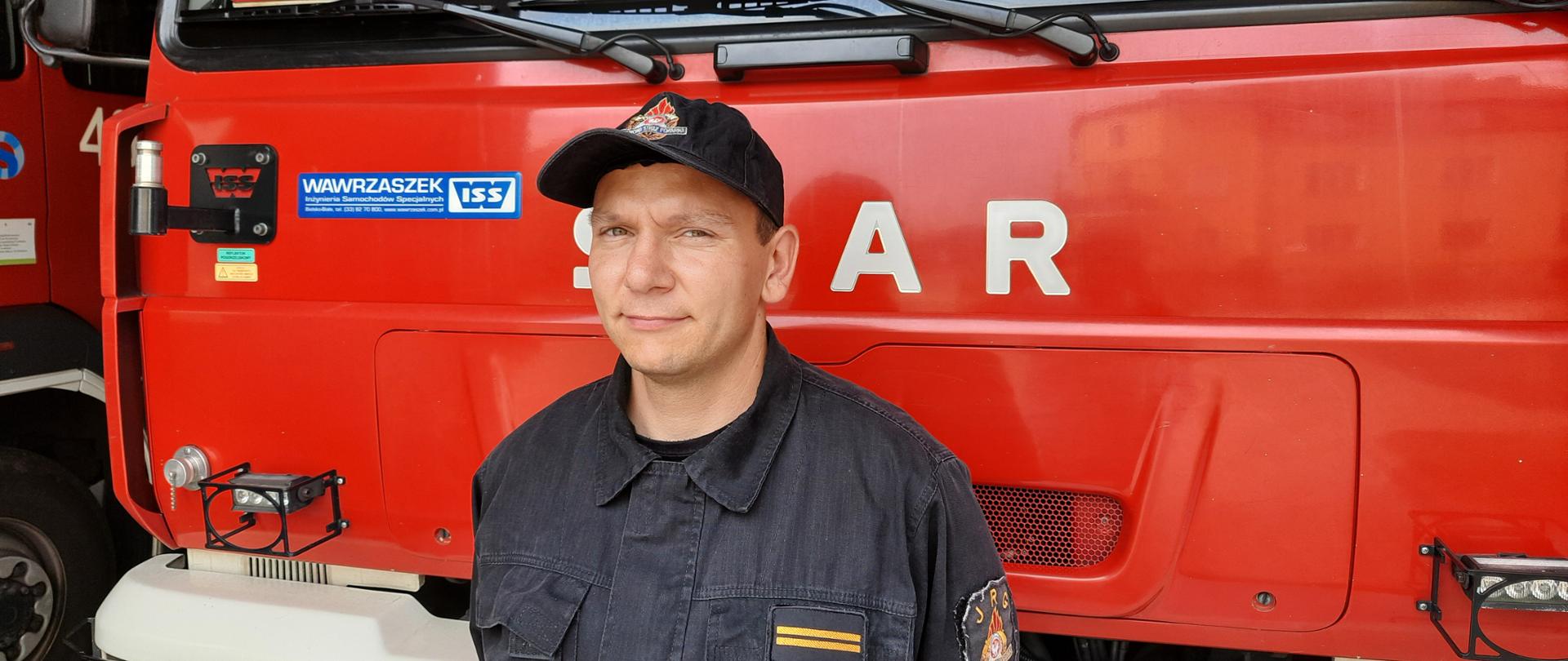 Zdjęcie przedstawia strażaka z Jednostki Ratowniczo-Gaśniczej Państwowej Straży Pożarnej w Lwówku Śląskim, który uratował kobietę podczas pożaru budynku. Strażak znajduje się na garażu jednostki na tle samochodu pożarniczego. 