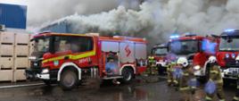 Zdjęcie przedstawia dynamiczną akcję gaśniczą na terenie przemysłowym. W tle widoczny jest fragment niebieskiej hali, zza której wydobywają się ogromne, kłębiaste chmury gęstego, szarego i białego dymu, przesłaniające niemal całe niebo.
Na pierwszym planie, po lewej stronie, stoi zaparkowany nowoczesny, czerwony wóz strażacki marki Scania. Pojazd ma otwartą jedną z bocznych rolet, odsłaniając specjalistyczny sprzęt ratowniczy. Podłoże jest mokre, asfaltowe, z widocznymi kałużami odbijającymi światła wozów.
Po prawej stronie zdjęcia znajduje się kilka innych czerwonych samochodów pożarniczych (w tym marki Scania oraz Iveco) z włączonymi niebieskimi sygnałami błyskowymi. Wokół pojazdów poruszają się strażacy w piaskowo-brązowych ubraniach specjalnych, hełmach oraz kamizelkach. Część z nich idzie w stronę przodu kadru, inni stoją w głębi, zabezpieczając miejsce zdarzenia.
Po lewej stronie kadru, obok niebieskiej hali, widać ułożone w stosy duże, jasne skrzyniopalety. Na całym zdjęciu widoczne są lekkie rozmycia i flary świetlne (efekt kropel wody lub dymu na obiektywie).