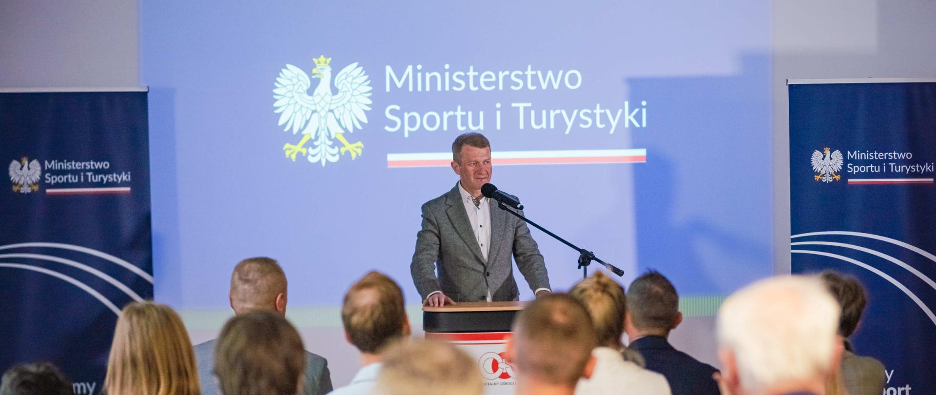 Minister Raś na konferencji trenerów stoi za mównica i mówi do mikrofonu. Na pierwszym planie słuchacze. Za plecami ministra ścianki z logotypami MSiT.