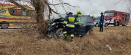 Czarny samochód osobowy wbity w drzewo na poboczy drogi. Wokół niego strażacy prowadzą działania. W tle karetka, samochód policyjny oraz wóz strażacki.
