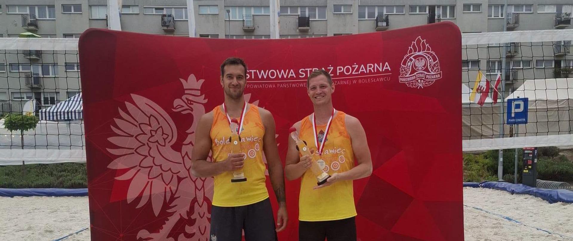 Zawodnicy KM PSP Wrocław stoją na tle baneru z napisem Państwowa Straż Pożarna z pucharami. 