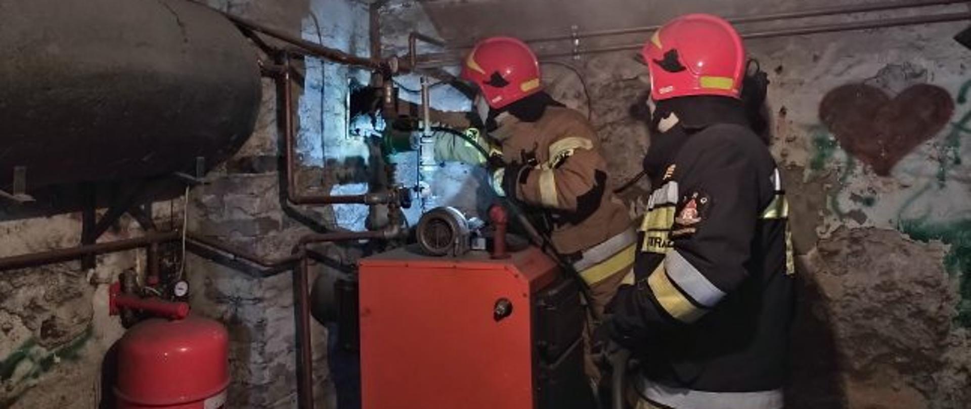 Na pierwszym planie dwóch strażaków sprawdzających przewód kominowy w kotłowni.
