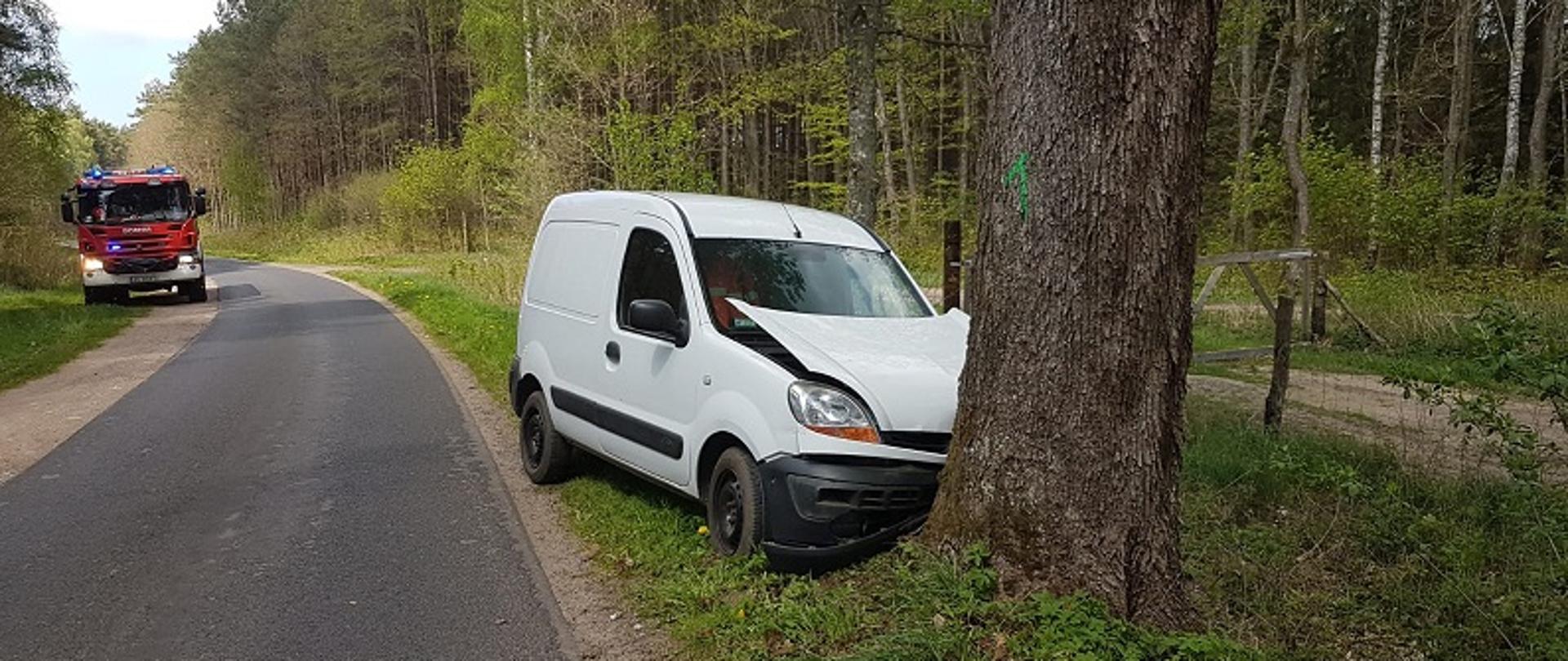 Zdjęcie przedstawia widoczny bokiem od strony pasażera uszkodzony pojazd marki Renault Kangoo. Samochód stoi na poboczu, przodem wbitym w pień drzewa. Za pojazdem na drodze samochód strażacki