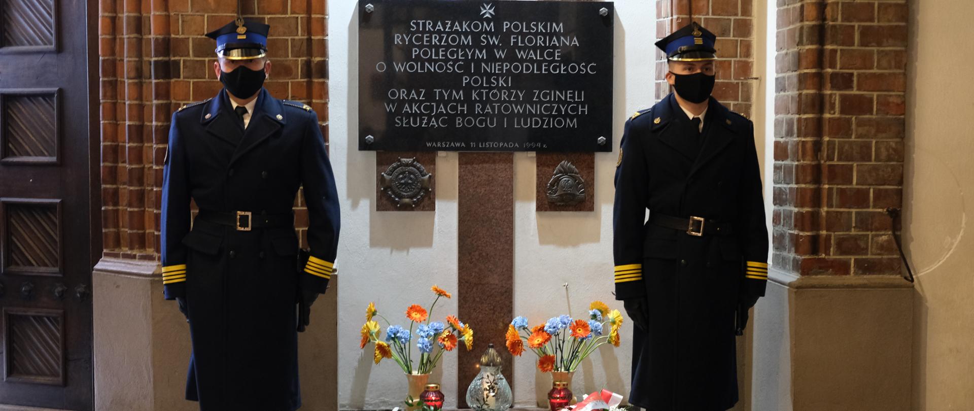 Dwóch strażaków stoi na baczność pełniąc wartę przy tablicy poległych strażaków