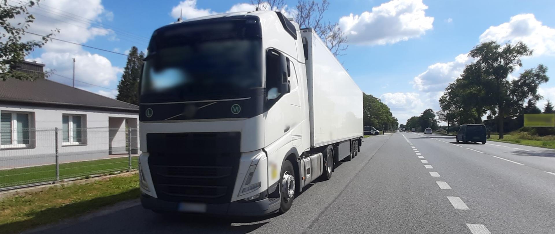 Na pierwszym planie (od lewej): przód i lewy bok ciągnika siodłowego z podpiętą naczepą - chłodnią. Zatrzymany do kontroli zestaw ciężarowy stoi na poboczu przy drodze krajowej. W tle: prawy bok i przód oznakowanego furgonu Inspekcji Transportu Drogowego i stojący obok niego umundurowany funkcjonariusz ITD.