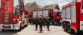Kompania honorowa PSP maszeruje pomiędzy samochodami strażackimi. Po bokach ustawione wozy bojowe i duży baner z napisem „Straż Pożarna”.
