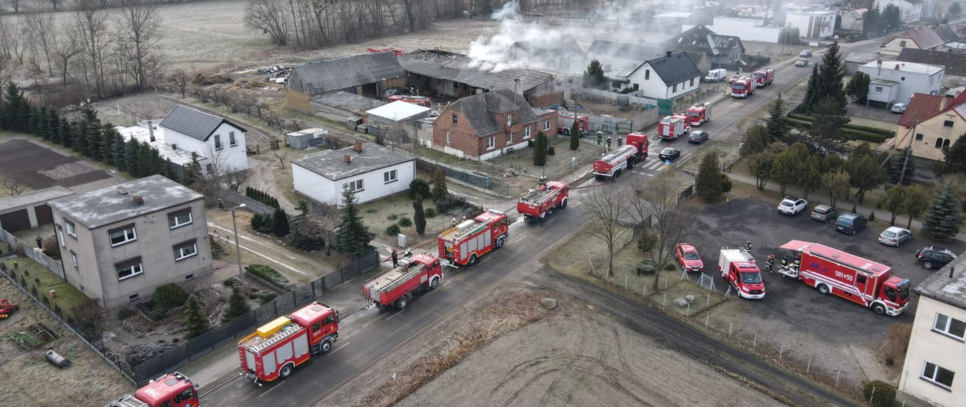 widok z góry ulica na której ustawione są samochody gaśnicze strażackie widoczne zabudowania z jednego z budynków wydobywa się biały dym