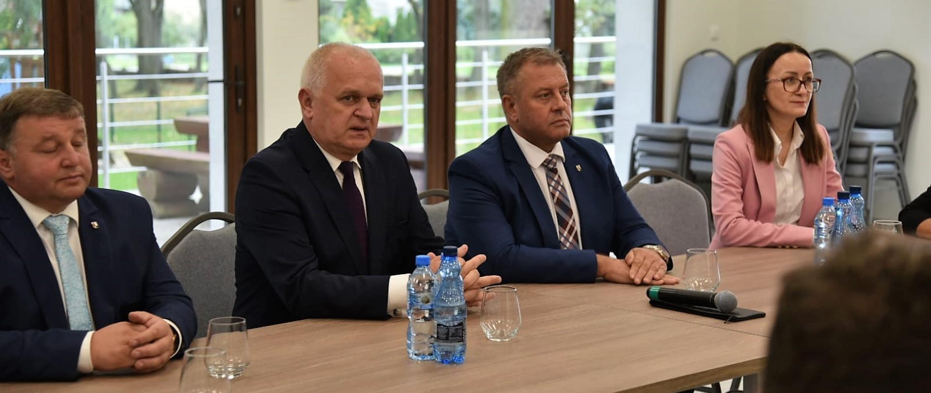 Wojewoda siedzi przy stole w jasno oświetlonej i przeszklonej sali. Po jego oby stronach siedzą samorządowcy 