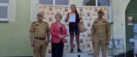 Fotografia przedstawia podium zwyciężczyni w kategorii kobiet wyścigu rowerowego MTB o puchar komendanta powiatowego. Po prawej i lewej stronie widać strażaków Państwowej Straży Pożarnej w ubraniu służbowym stojących obok podium. Wygrana prezentuje dyplomy oraz statuetki.