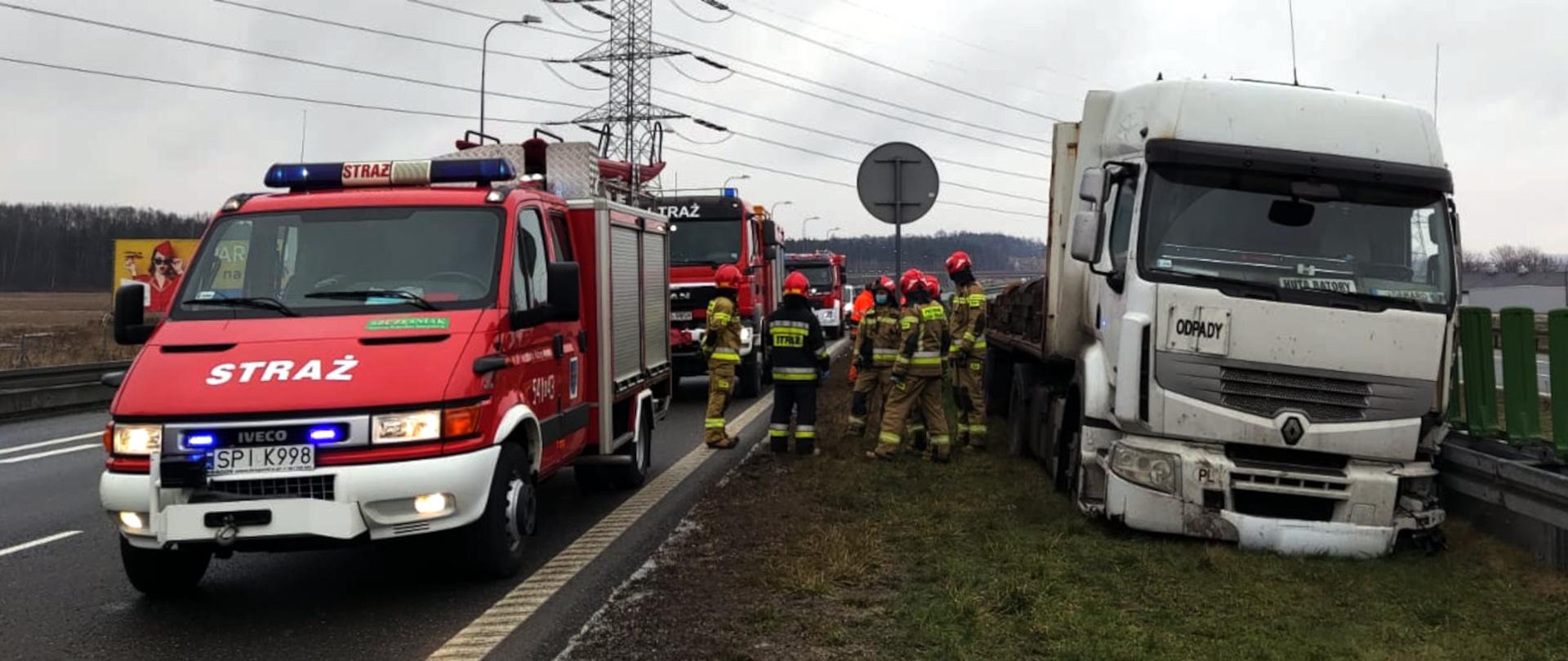 Zdjęcie przedstawia z lewej strony trzy samochody pożarnicze z włączonymi światłami ostrzegawczymi stojące na pasie ruchu autostrady, z lewej strony samochód ciężarowy stojący na pasie zieleni i oparty o barierę energochłonną. Na środku widać grupę strażaków w trakcie czynności ratowniczych.