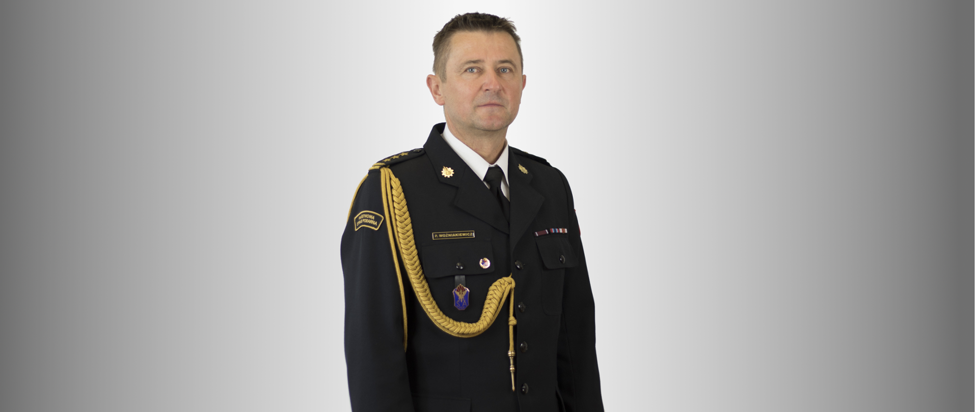 Na zdjęciu stoi mężczyzna w wieku około pięćdziesięciu lat, sfotografowany od kolan w górę, na jasnoszarym tle. Jest zwrócony lekko bokiem, ale patrzy prosto w obiektyw. Ma krótkie, ciemne włosy z lekką siwizną po bokach i spokojny, neutralny wyraz twarzy.
Ubrany jest w ciemny, elegancki mundur galowy Państwowej Straży Pożarnej. Mundur jest starannie zapięty, z białą koszulą i czarnym krawatem pod spodem. Na ramionach i kołnierzu znajdują się złote oznaczenia stopnia. Przez prawe ramię biegnie gruby, ozdobny złoty sznur, który opada na przód munduru. Na lewej piersi widoczne są drobne kolorowe wstążki odznaczeń i przypinki.
Na prawym rękawie znajduje się ciemna naszywka z napisem „Państwowa Straż Pożarna”. Całość wygląda bardzo oficjalnie — jak portret służbowy wysokiego rangą strażaka lub dowódcy