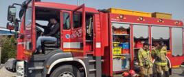 Na zdjęciu widać pojazd ratowniczo-gaśniczy na podwoziu MAN. Pojazd w kolorze czerwonym z oznaczeniami i sygnalizacją ostrzegawczą. Zdjęcie wykonane w ciągu dnia. Słonecznie. Na zdjęciu osoby strażacy ubrani w ubrania specjalne.