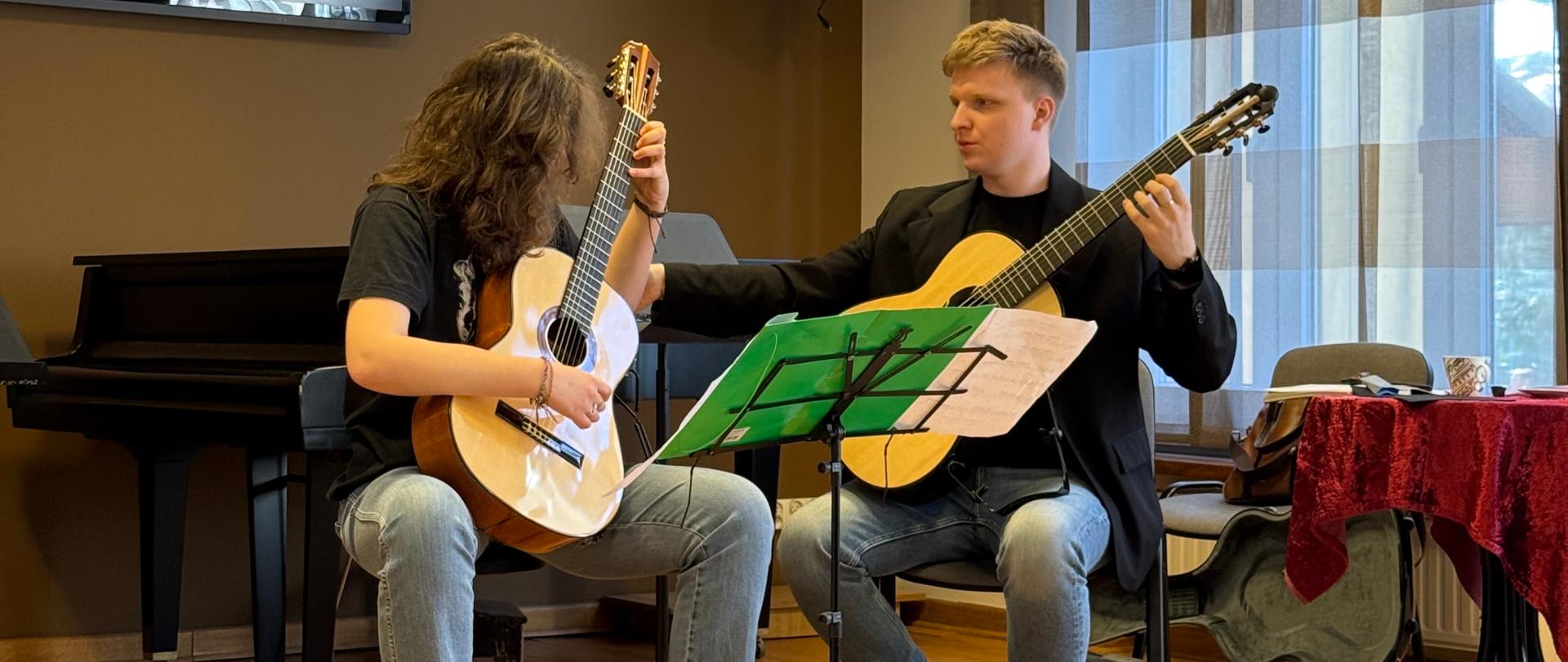 Na zdjęciu widnieje uczennica Aldona Gorczyca oraz prowadzący warsztaty mgr Mateusz Kowalski który instruuje uczennice w trakcie lekcji mistrzowskiej. Kolorystyka zdjęcia jest żółto-beżowo-brązowo-biało-niebiesko-czerwono-czarna.