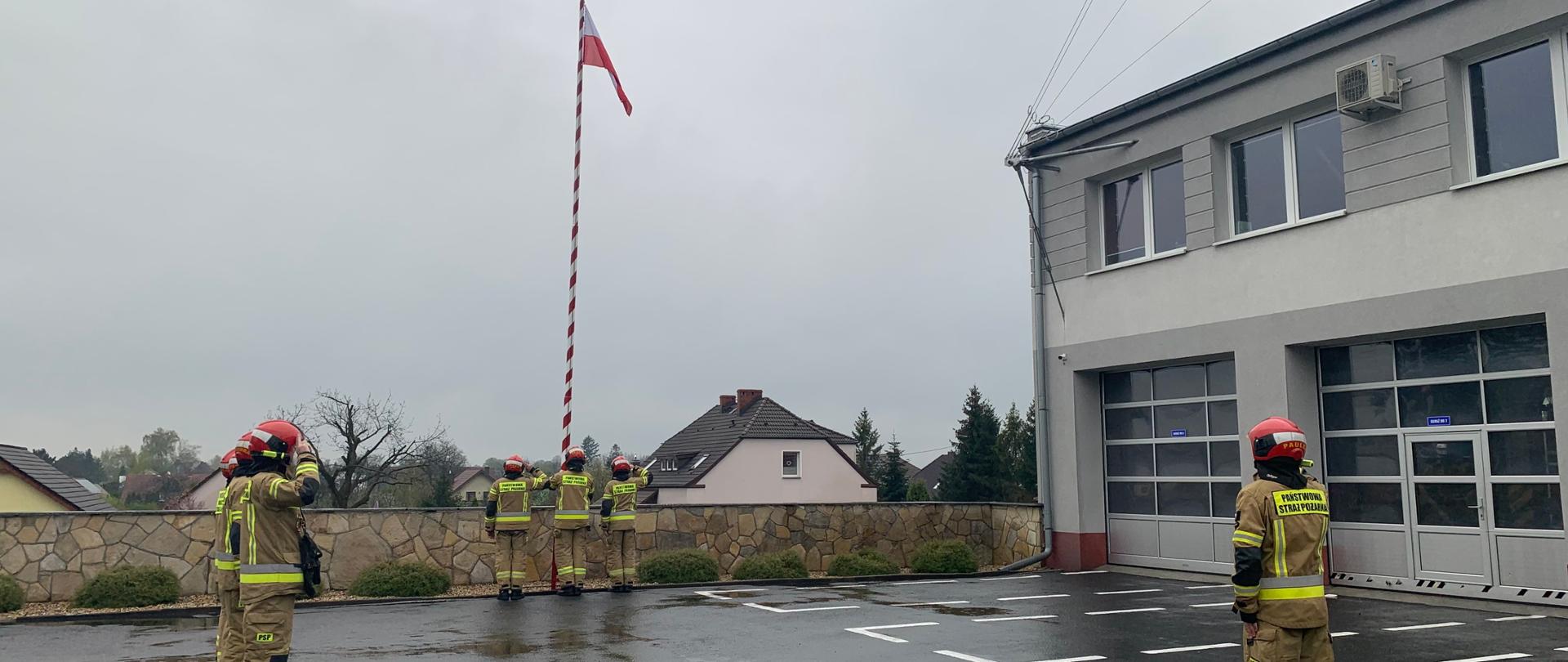 Uroczysta zmiana służby z okazji Święta Flagi RP. W centrum zdjęcia, na biało-czerwony maszt poczet flagowy podnosi polską flagę narodową. Po lewej stronie widać strażaków zmiany służbowej. po prawej dowódca uroczystości. Dowódca zmiany oraz dowódca pododdziału a także dwaj strażacy w poczcie salutują. Strażacy w ubraniach specjalnych koloru pisakowego i czerwonych hełmach.