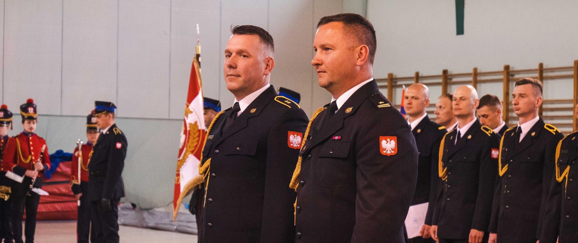 dwoch strażakó stoi obok sienie w tle grupa strażaków w szeregu oraz poczet sztandarowy