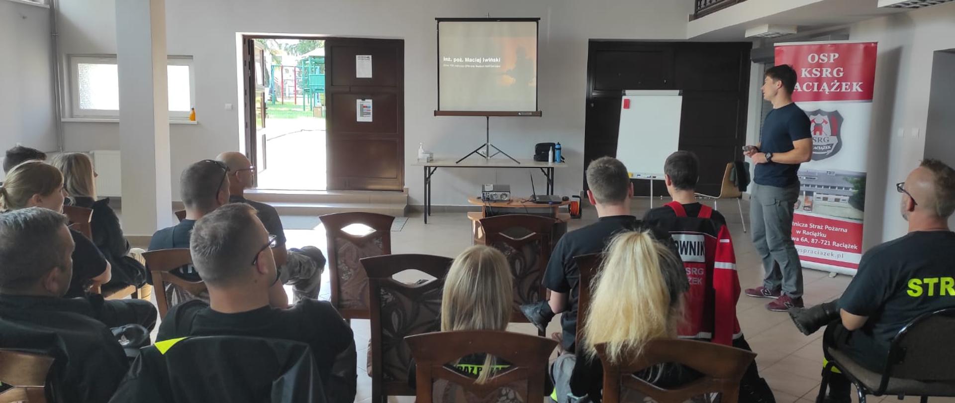 Zdjęcie obrazuje siedzących na drewnianych krzesłach wyścielonych tapicerką tekstylną strażaków tyłem do wykonującego fotografię. Zdjęcia zrobiono na Sali wykładowej w budynku ochotniczej straży pożarnej w Raciążku, ściany pomalowane na jasny kolor. W tle zdjęcia widać dwie pary brązowych drzwi dwuskrzydłowych, gdzie, jedne z nich są otwarte. W ścianie z drzwiami powyżej zamontowane są okna. Pomiędzy wspomnianymi drzwiami stoi drewniany stół na czarnych metalowych nogach. Na stole stoi ekran, na którym, rzutnik wyświetla tematykę związaną z prowadzeniem działań gaśniczych podczas pożarów. Widać łącznie dwanaście osób biorących udział w szkoleniu ubrani są w umundurowanie koszarowe koloru czarnego. Wśród tych dwunastu osób są cztery kobiety. Po prawej stronie zdjęcia stoi mężczyzna ubrany w czarną koszulkę na krótki rękaw oraz siwe długie spodnie i wiśniowe sportowe buty widać, iż omawia on temat wyświetlany na ekranie.