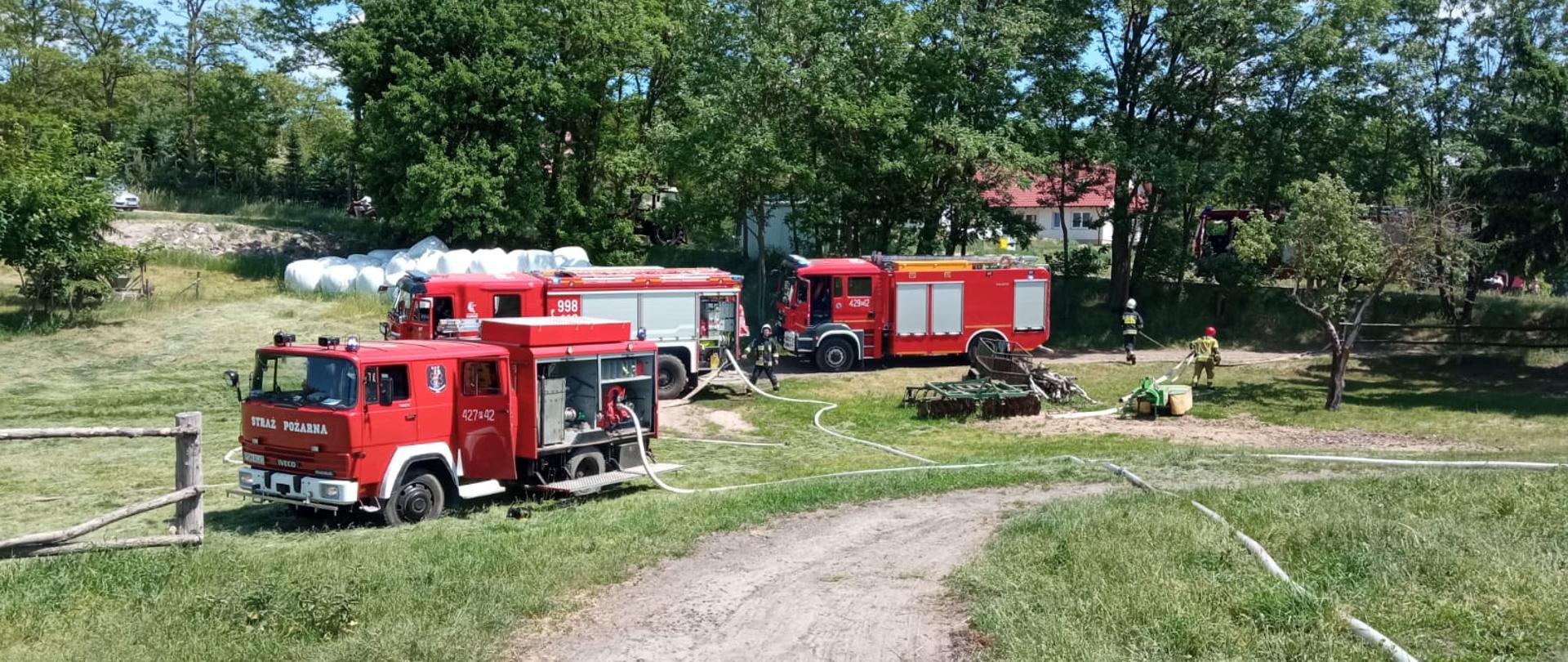 Na zdjęciu widać trzy samochody pożarnicze od których rozwinięte są węże. Jest pogodny słoneczny dzień wokół samochodów jest dużo zieleni. Z tył za samochodami składowane są baloty