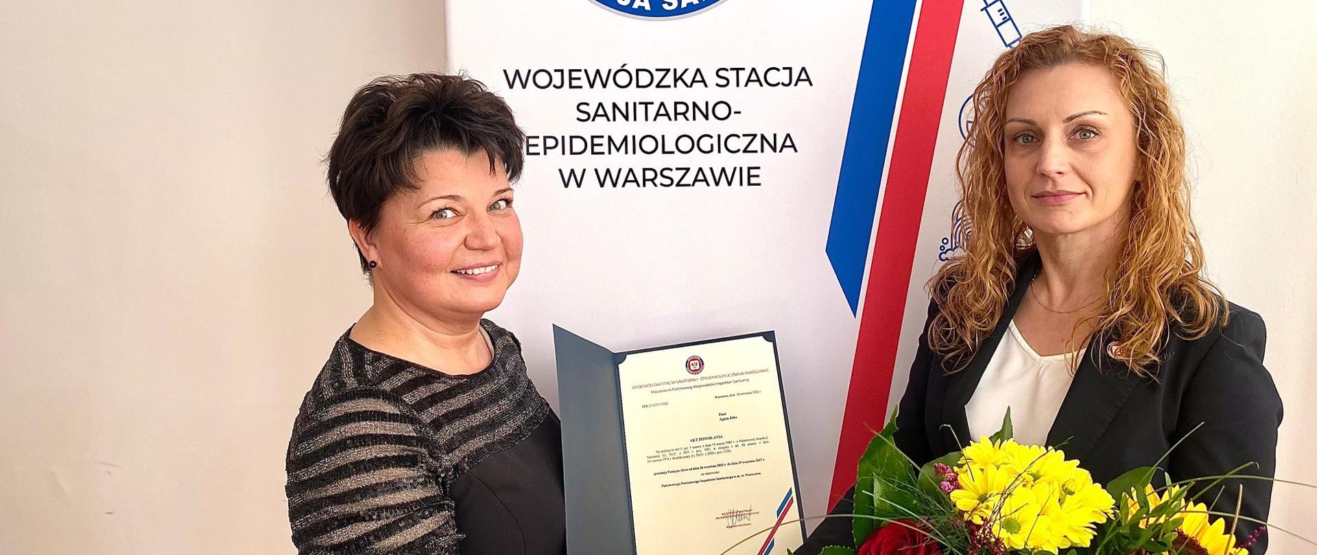 dwie kobiety na tle logo PIS, kobieta po lewej trzyma dokument z powołaniem na PPIS w m.st. Warszawie, kobieta po prawej trzyma bukiet kwiatów i prawą ręką ściska dłoń kobiety po lewej stronie, gratulując przyjętej nominacji