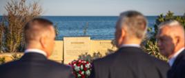 Na zdjęciu widać trzech mężczyzn stojących tyłem do obiektywu (w środku minister Tomasz Siemoniak). Mężczyźni stoją przed nagrobkiem polskiego inżyniera Pawła Potockiego. Nagrobek składa się z trzech tablic w różnych językach. Pod środkową tablicą w języku polskim leży wieniec w kolorach czerwonym (róże), białym (gerbery) i zielonym z biało-czerwoną szarfą. Po prawej i lewej stronie tablic nagrobkowych widać roślinność, a w tle niebieskie morze. 